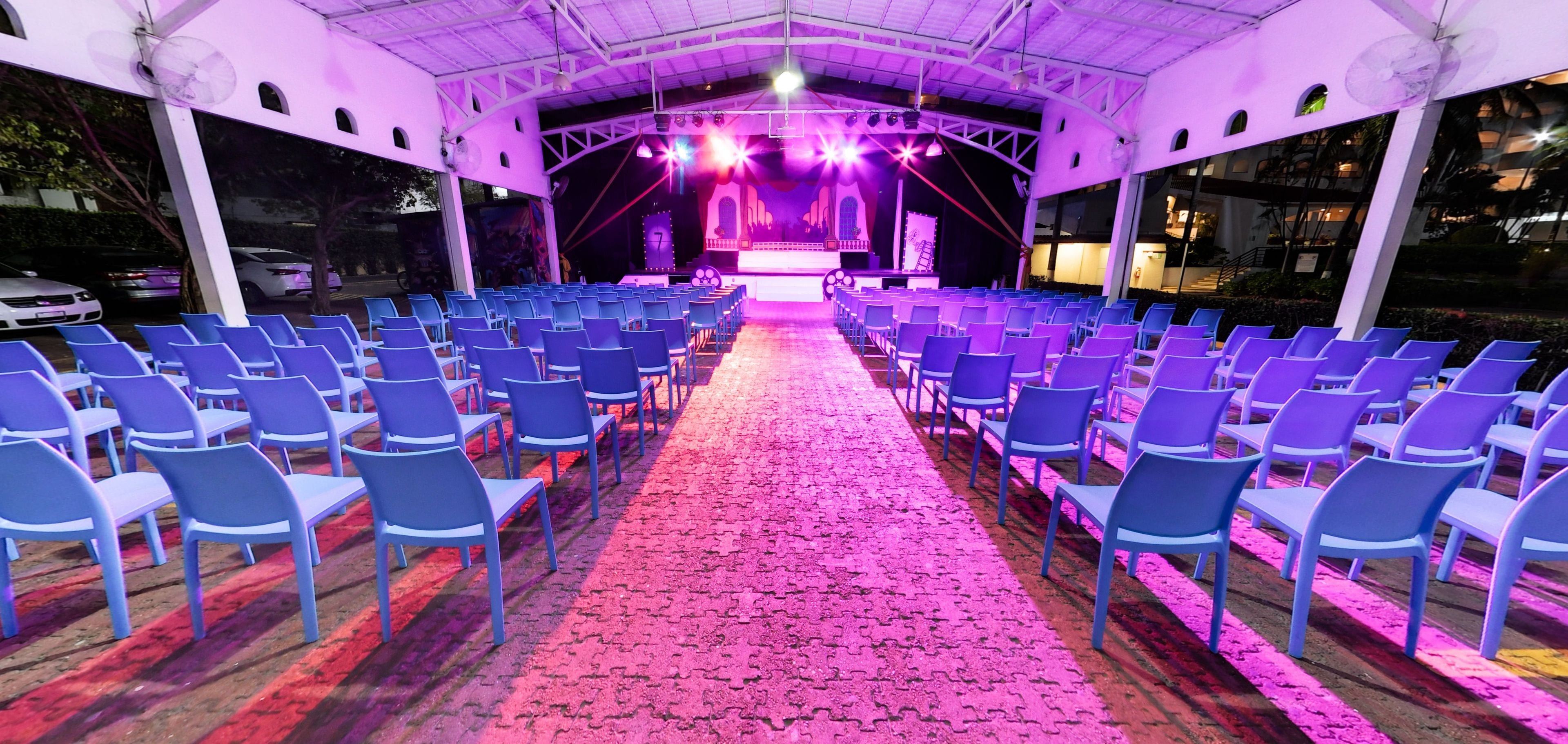Espaço interior com fileiras de cadeiras azuis e palco iluminado em roxo.