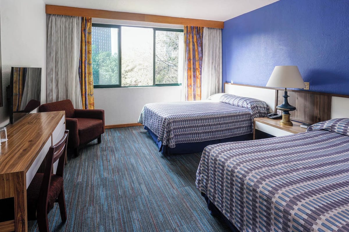 A hotel room with two beds, a desk, a chair, a lamp, and a view of trees through the window.