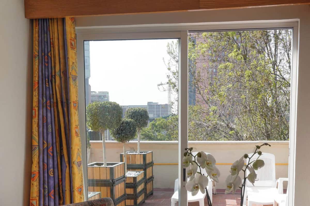 A room with a patterned armchair, a small table with a plant, and a view of a patio with outdoor furniture and trees.