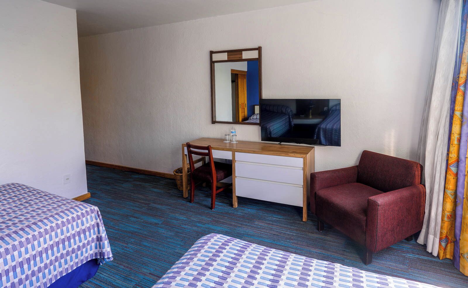 Quarto de hotel com duas camas de solteiro, uma mesa com cadeira, televisão e uma poltrona vermelha.