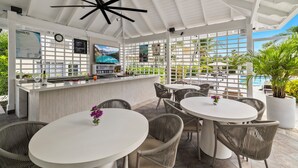 A covered outdoor seating area with white tables and wicker chairs, a bar with a television, and a view of a pool. - Ports of Call Resort Turks, Tapestry Collection by Hilton (Providenciales)