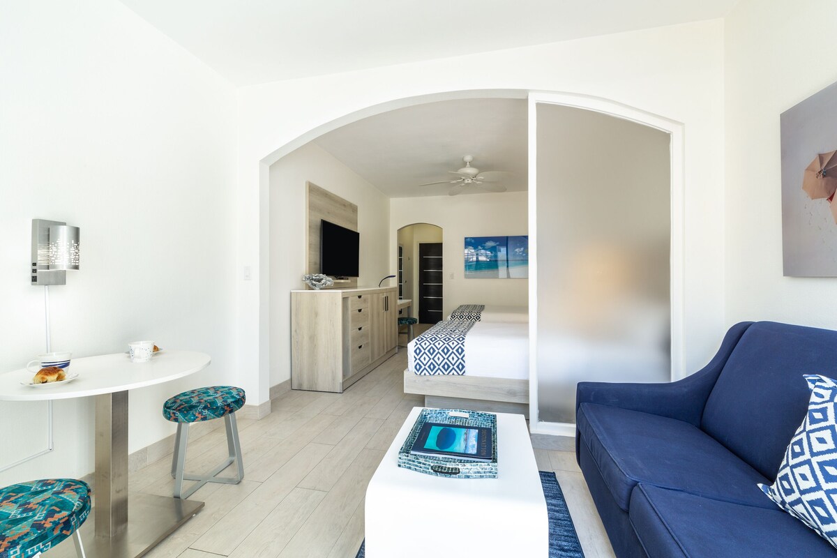 Een moderne woonkamer met een blauwe bank, een witte salontafel en een keukenhoek met een houten kast en een flatscreen televisie.