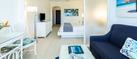 A modern hotel room with a dining area, a sofa, a ceiling fan, a bed with a patterned bedspread, and a painting of a beach scene.