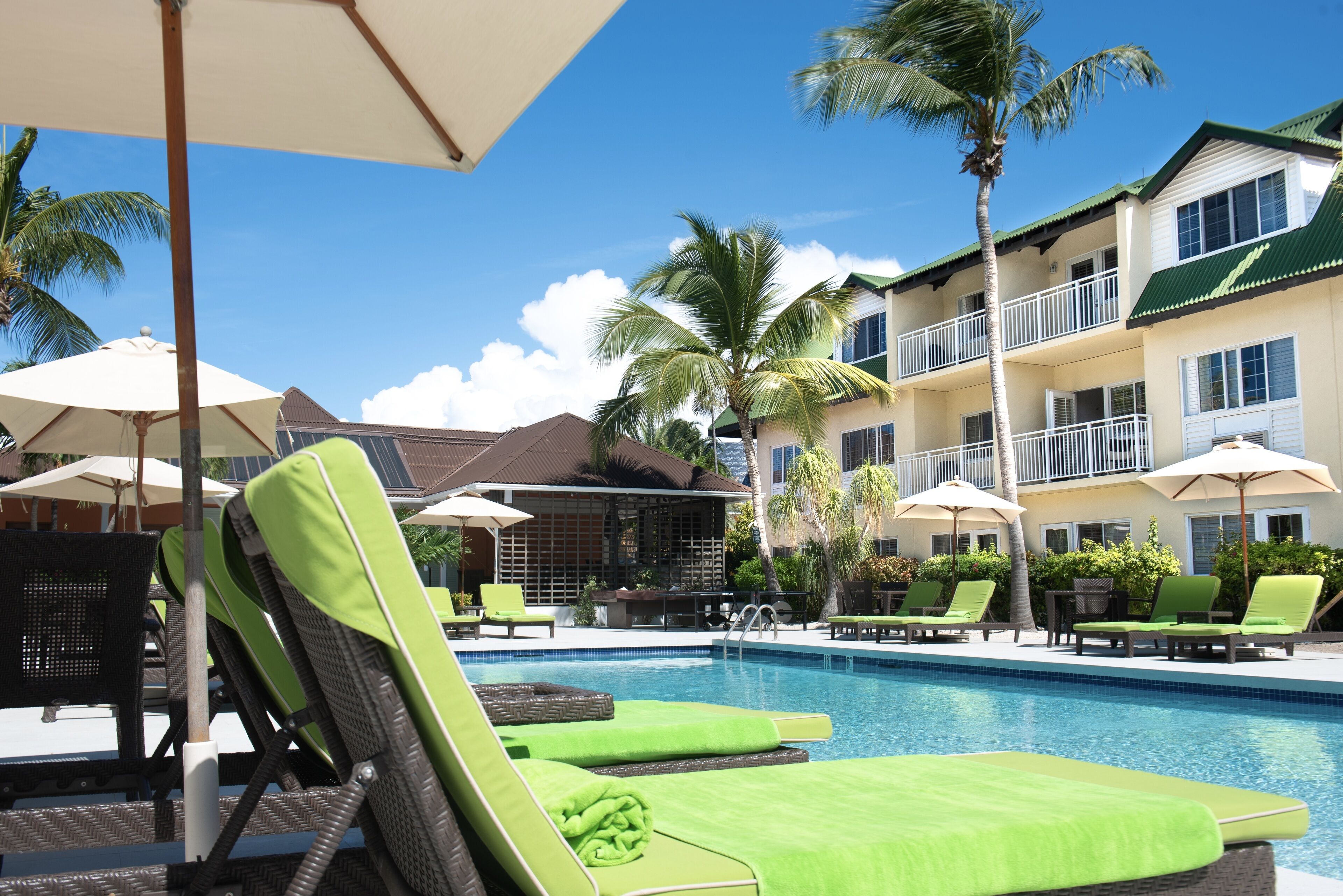 A poolside area with green lounge chairs and umbrellas, a clear blue sky, and a building with balconies and palm trees.