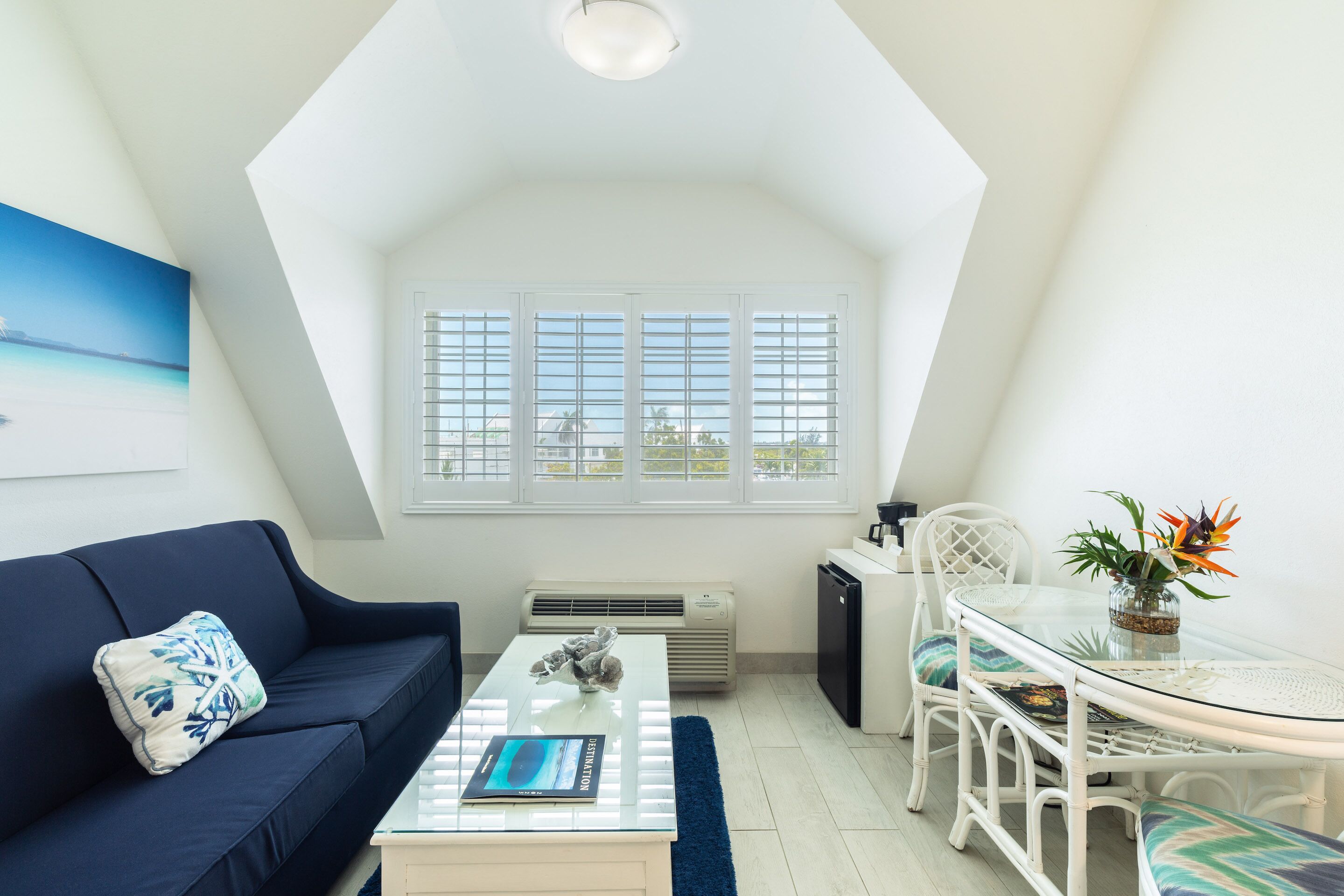 Une pièce moderne et bien éclairée, avec un canapé bleu, une table basse en verre, une table à manger avec des chaises et une grande fenêtre avec des volets.