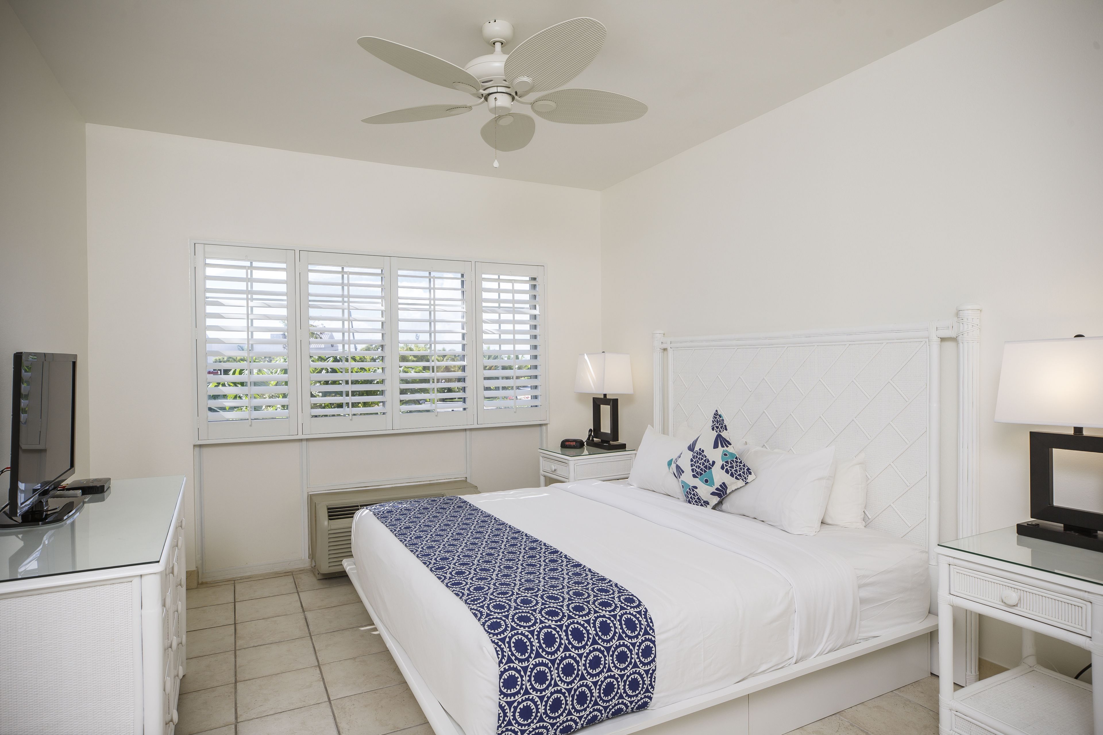 A bedroom with a bed, a ceiling fan, a TV on a desk, and a window with blinds.