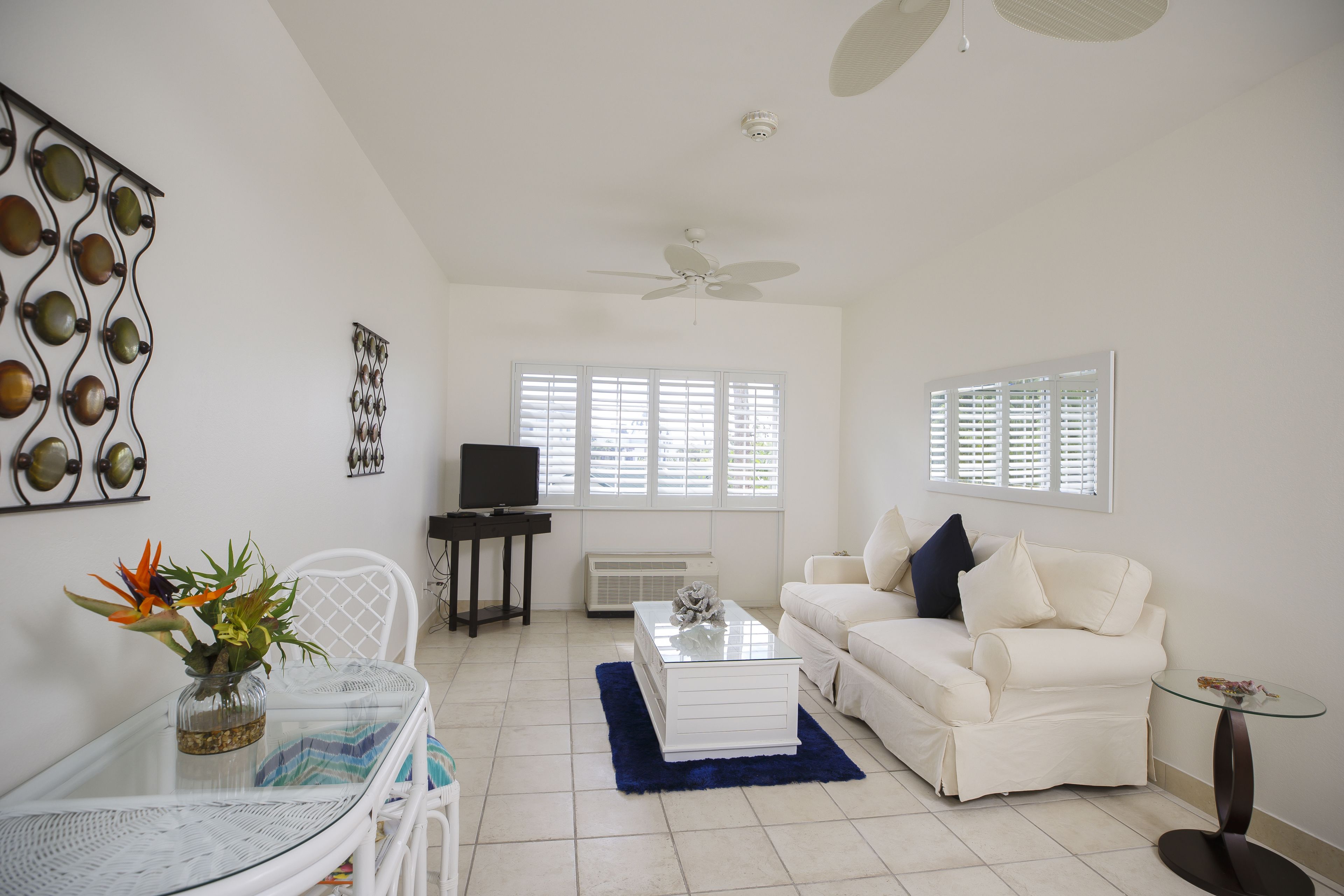 A living room with a white sofa, a glass coffee table, a white TV stand, and a ceiling fan.