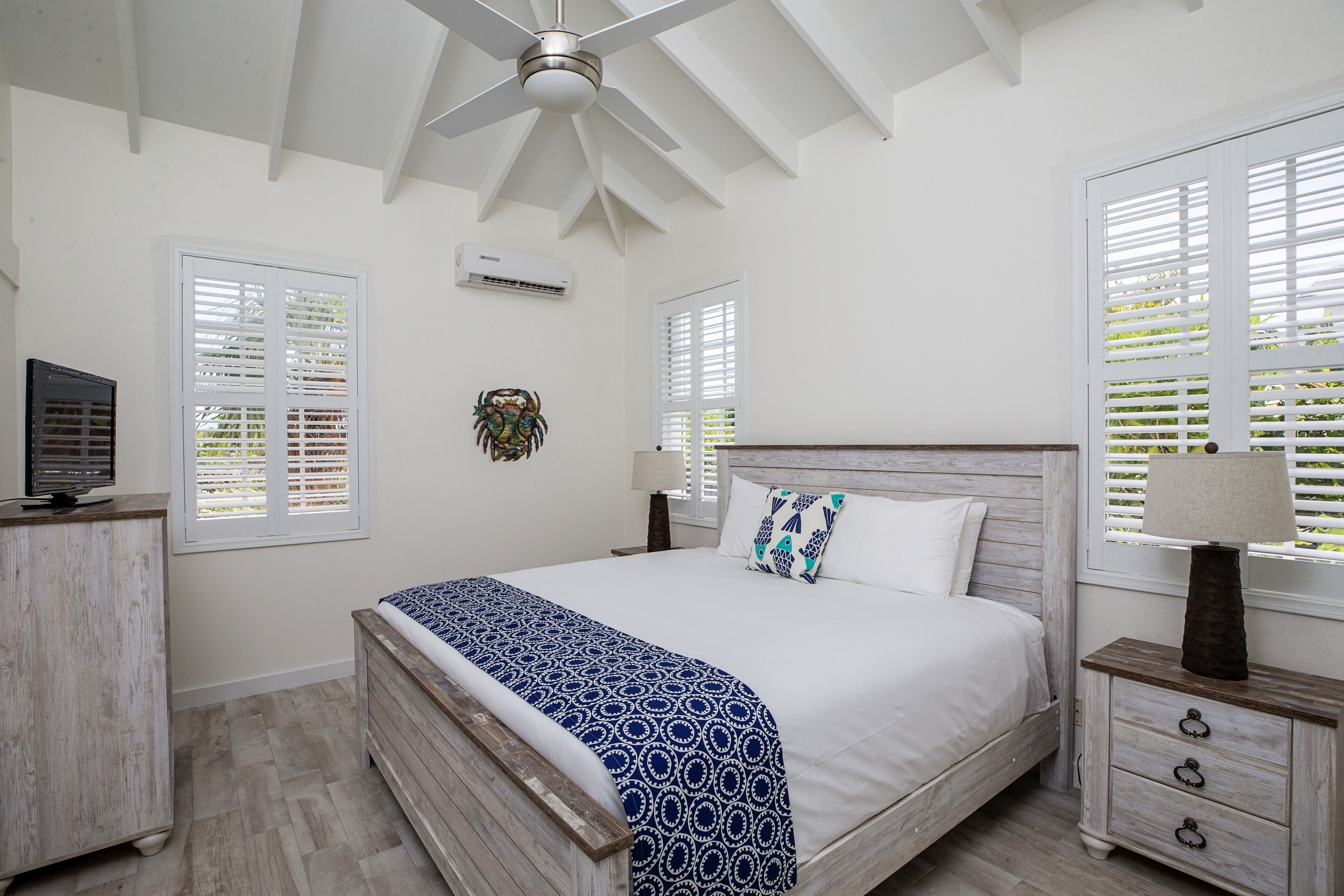 A bedroom with a bed, bedside tables, a ceiling fan, and windows with shutters.