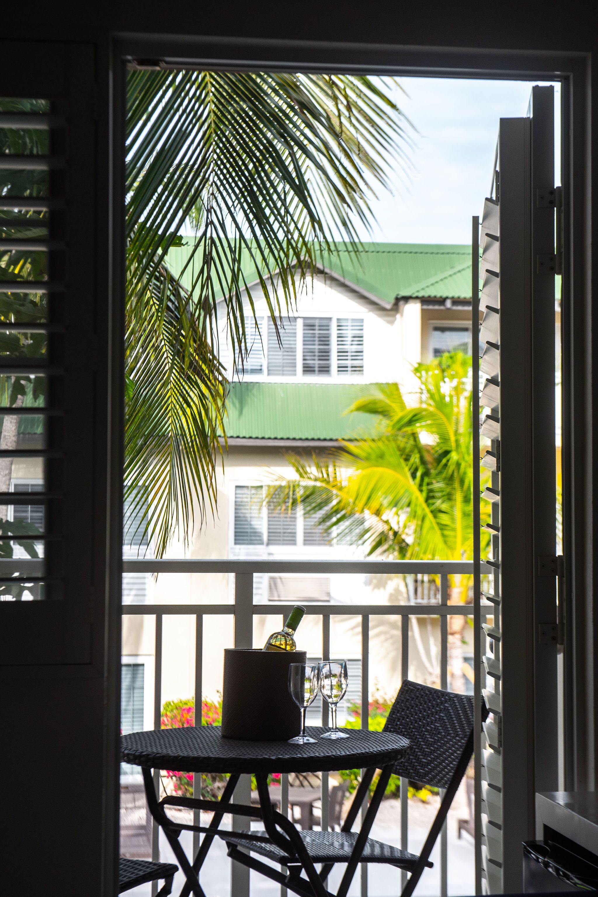 A balcony with a table and chairs, a potted plant, and a view of a building with a green roof and palm trees.