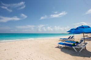 A beach with white sand, clear turquoise water, and blue umbrellas providing shade. - Ports of Call Resort Turks, Tapestry Collection by Hilton (Providenciales)