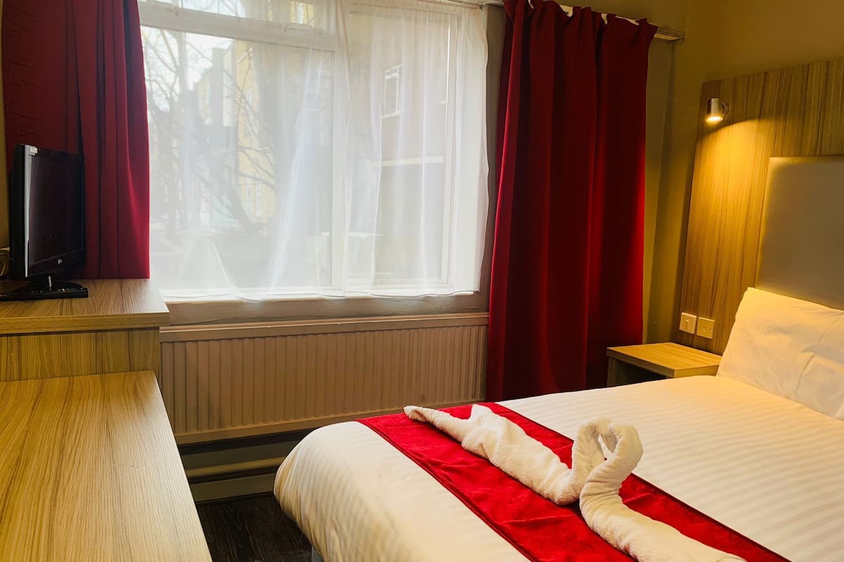 A hotel room with a bed, a desk with a television, a window with curtains, and a bedside lamp.