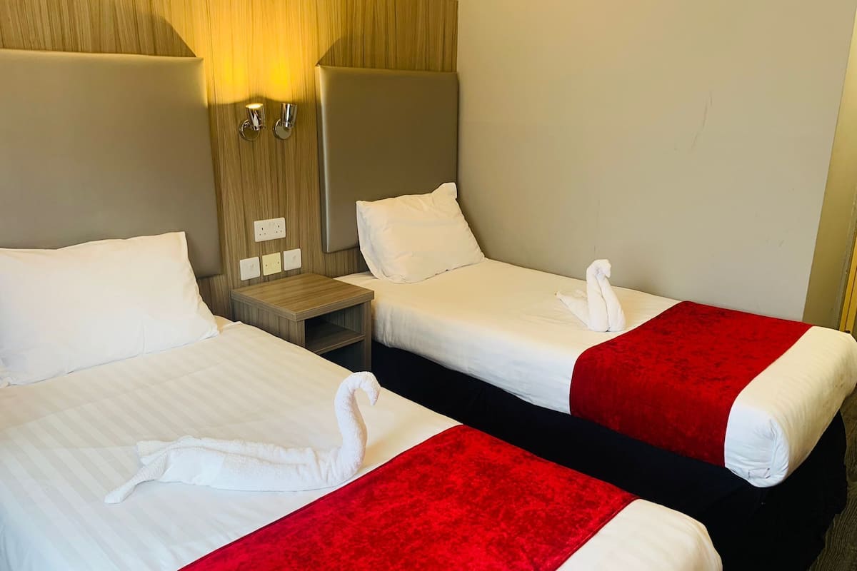 A hotel room with two beds, each with white linens and red throw pillows. There are wall-mounted lights and a wooden headboard.