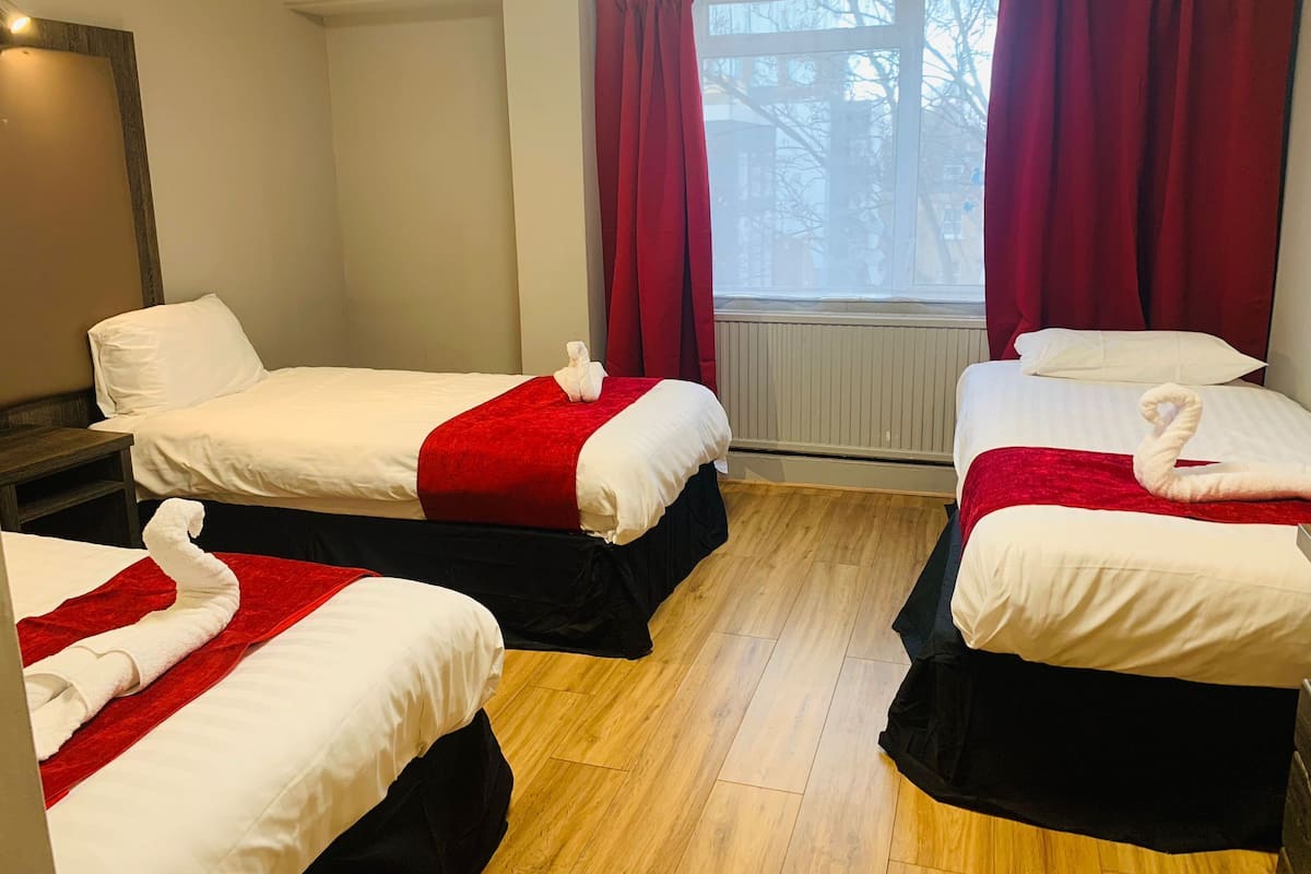A hotel room with two beds, each with a red blanket and a white towel shaped like a swan. The room has wooden flooring, a window with red curtains, and a radiator.