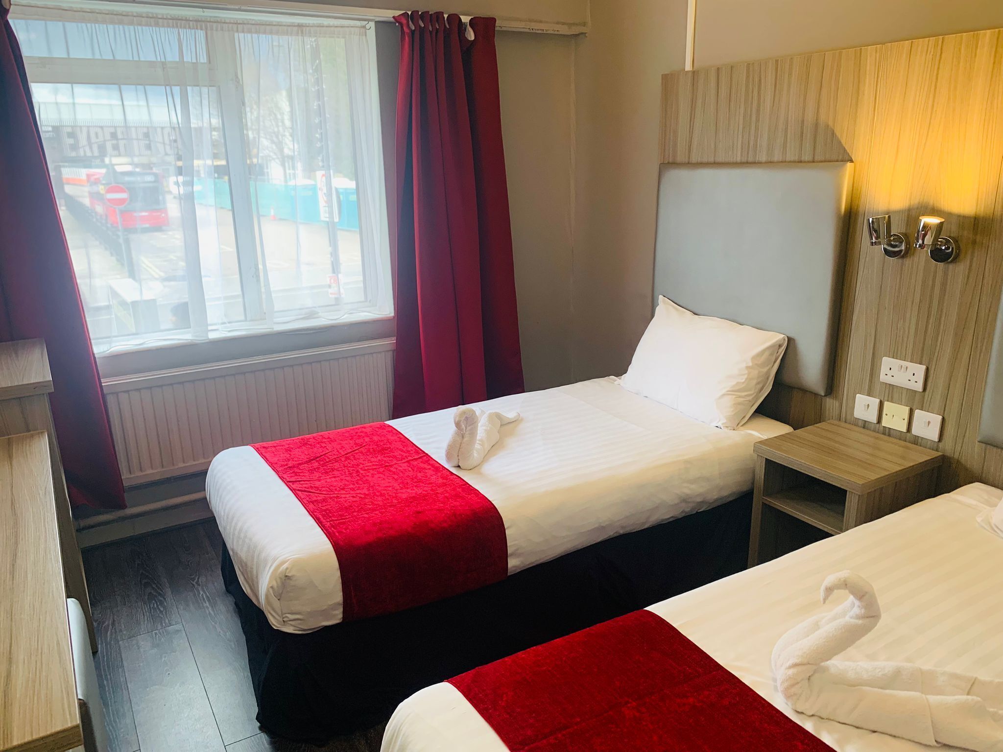 Ein Hotelzimmer mit zwei Betten, einem Fenster mit Vorhängen, einem Nachttisch mit Lampe und einem an der Wand montierten Lichtschalter.