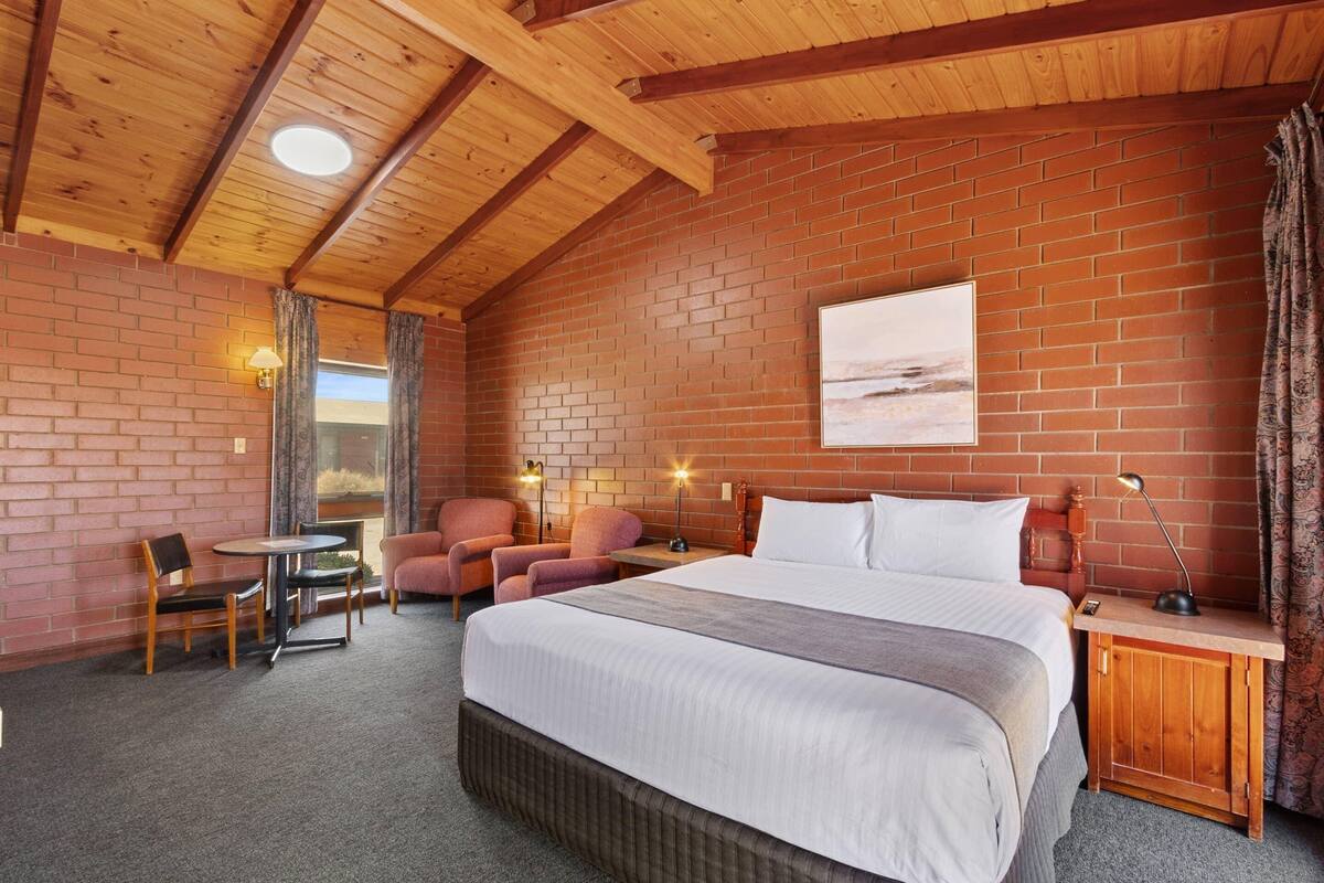 A hotel room with a large bed, a wooden ceiling, brick walls, a small table, two chairs, and a nightstand with a lamp.