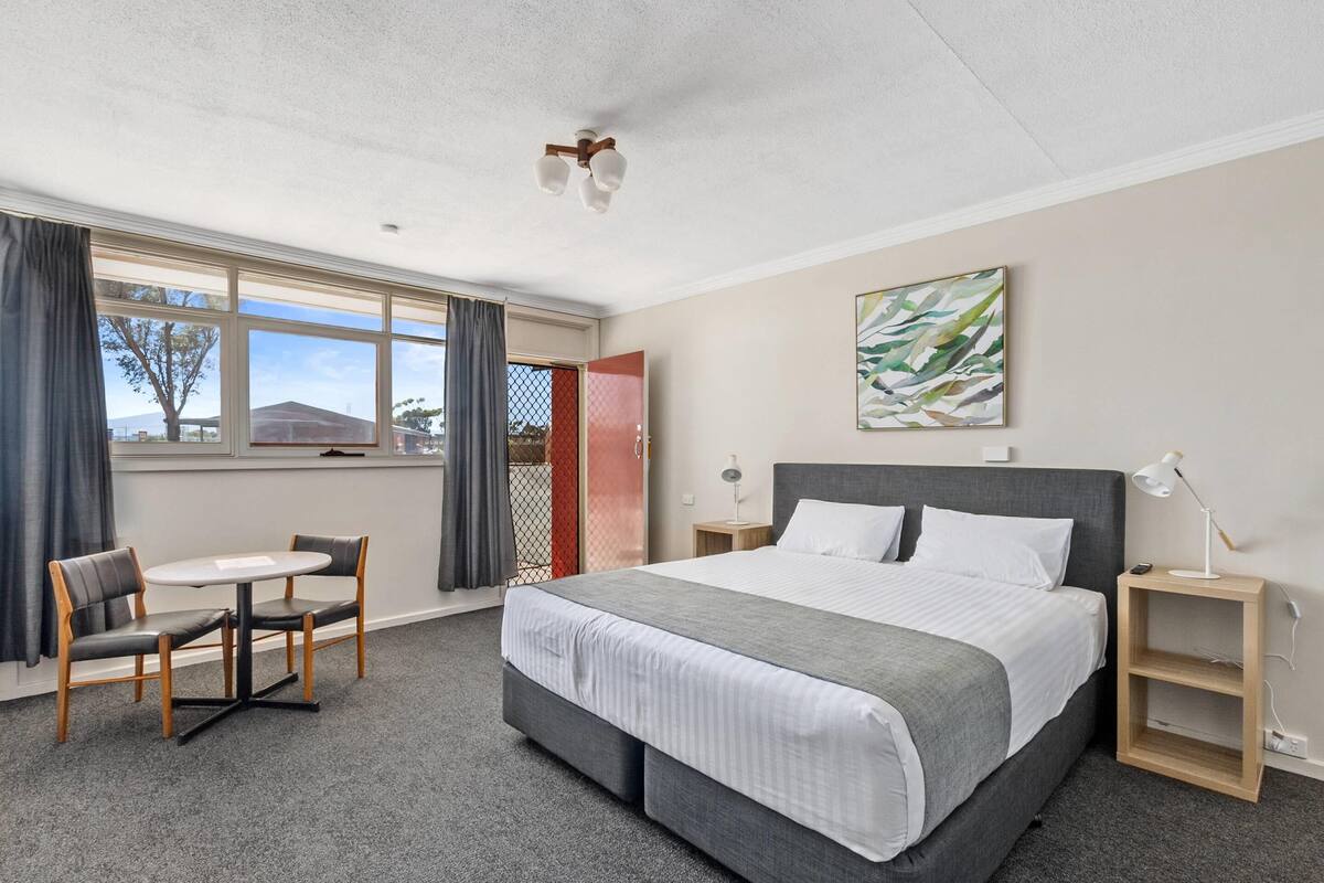 A hotel room with a large bed, a desk with two chairs, a window with curtains, a ceiling fan, and a painting on the wall.
