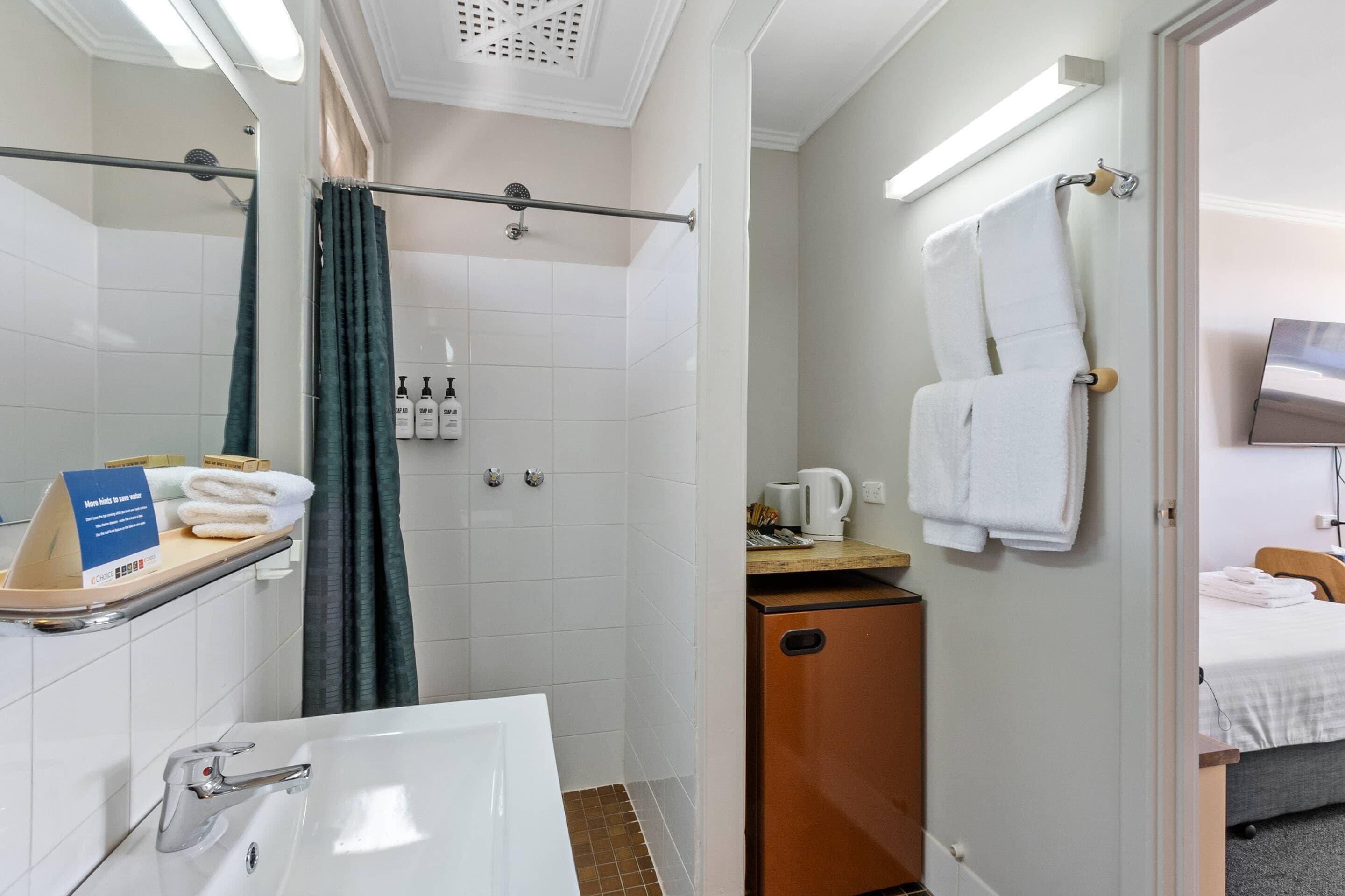 A hotel bathroom with a shower, a sink, and a toilet. There is a towel rack with towels, a small cabinet, and a sign indicating the room's amenities.