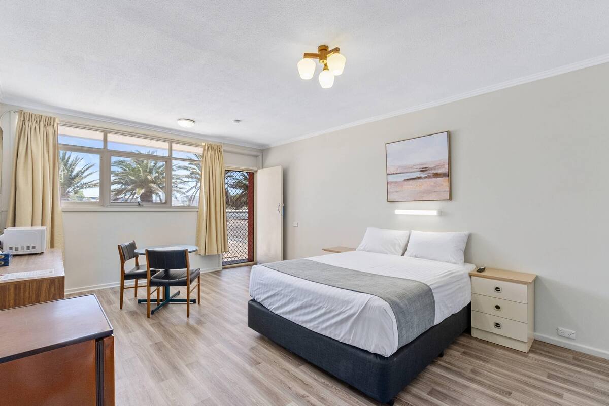 A hotel room with a bed, bedside table, desk, chair, and a view of palm trees through the window.