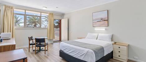 A hotel room with a bed, bedside table, desk, chair, and a view of palm trees through the window.