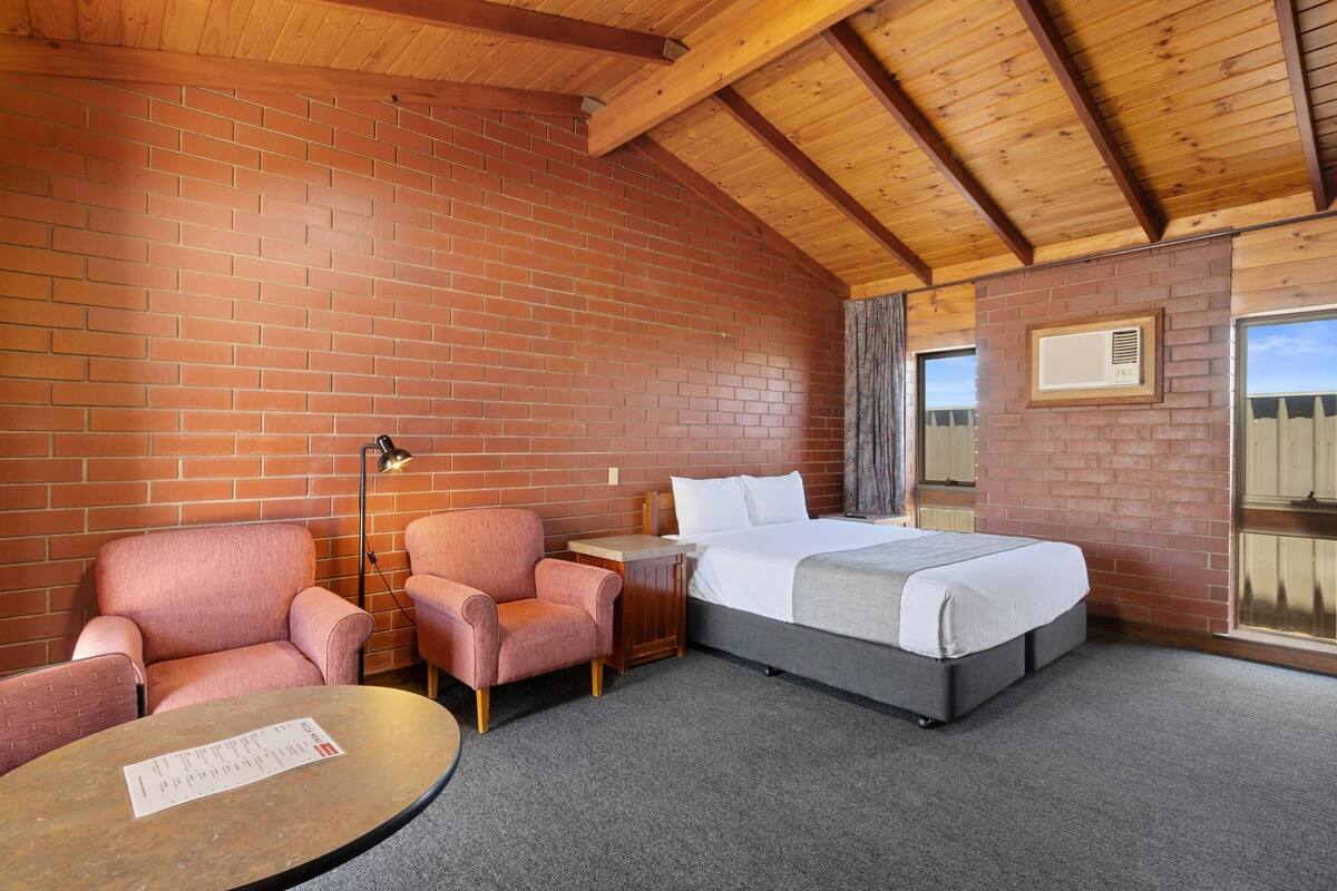 A room with a brick wall, wooden ceiling beams, a bed, two pink armchairs, a small table with a menu, and a window with a view.