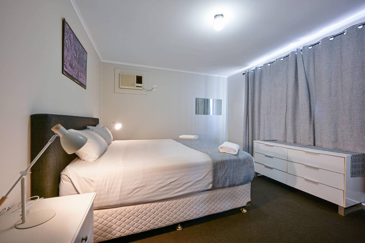 A hotel room with a bed, bedside lamp, dresser, and a window with curtains.