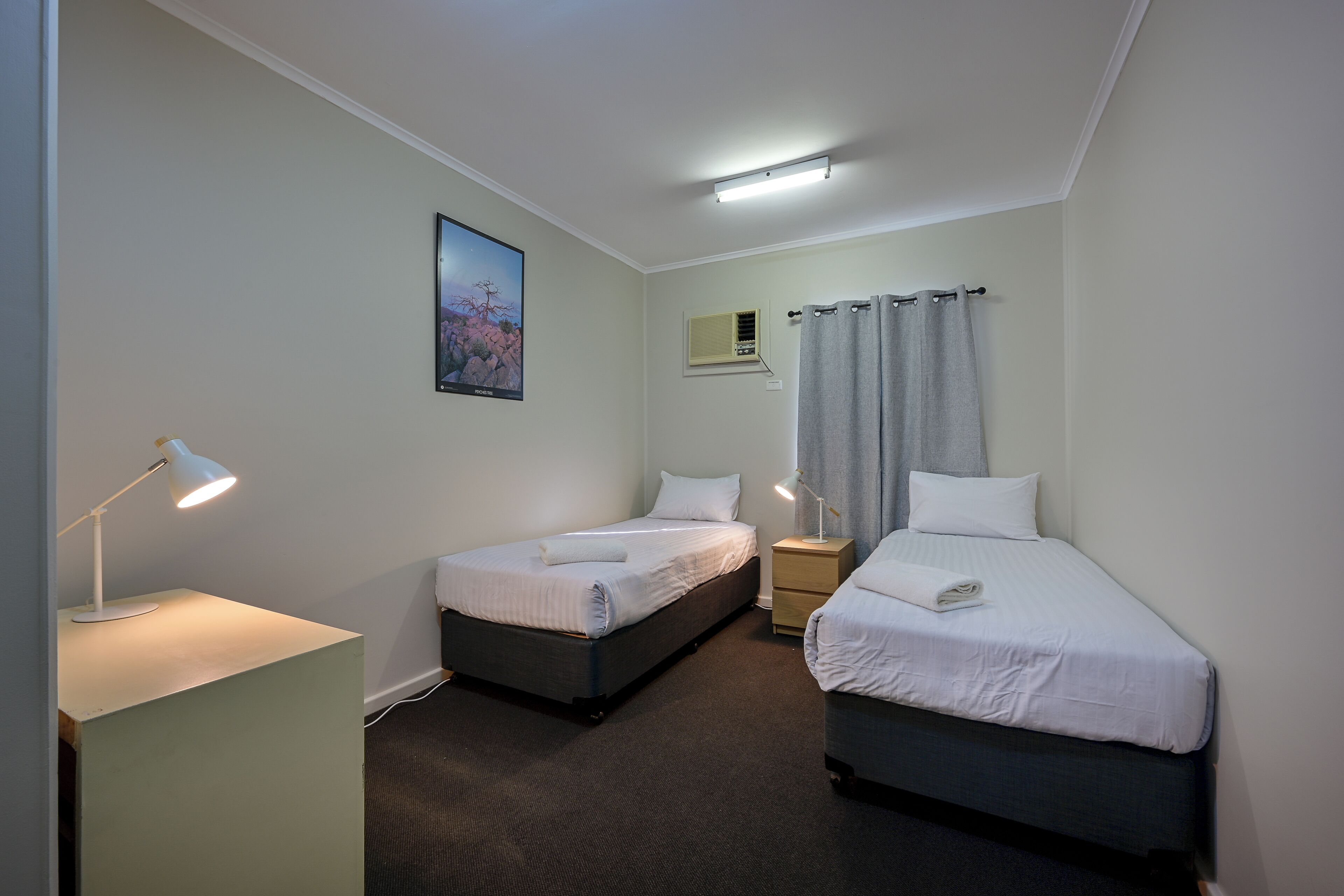 A hotel room with two single beds, a desk with a lamp, a small bedside table, a wall-mounted air conditioning unit, and a framed picture on the wall.