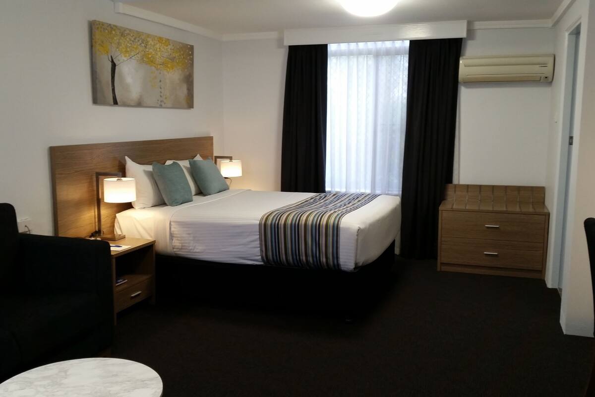 A hotel room with a bed, bedside tables, a desk, a chair, and a painting on the wall.