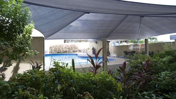 A covered outdoor area with a swimming pool, surrounded by greenery and a shaded canopy. - Mid City Motor Inn (Singleton)