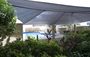 A covered outdoor area with a swimming pool, surrounded by greenery and a shaded canopy.