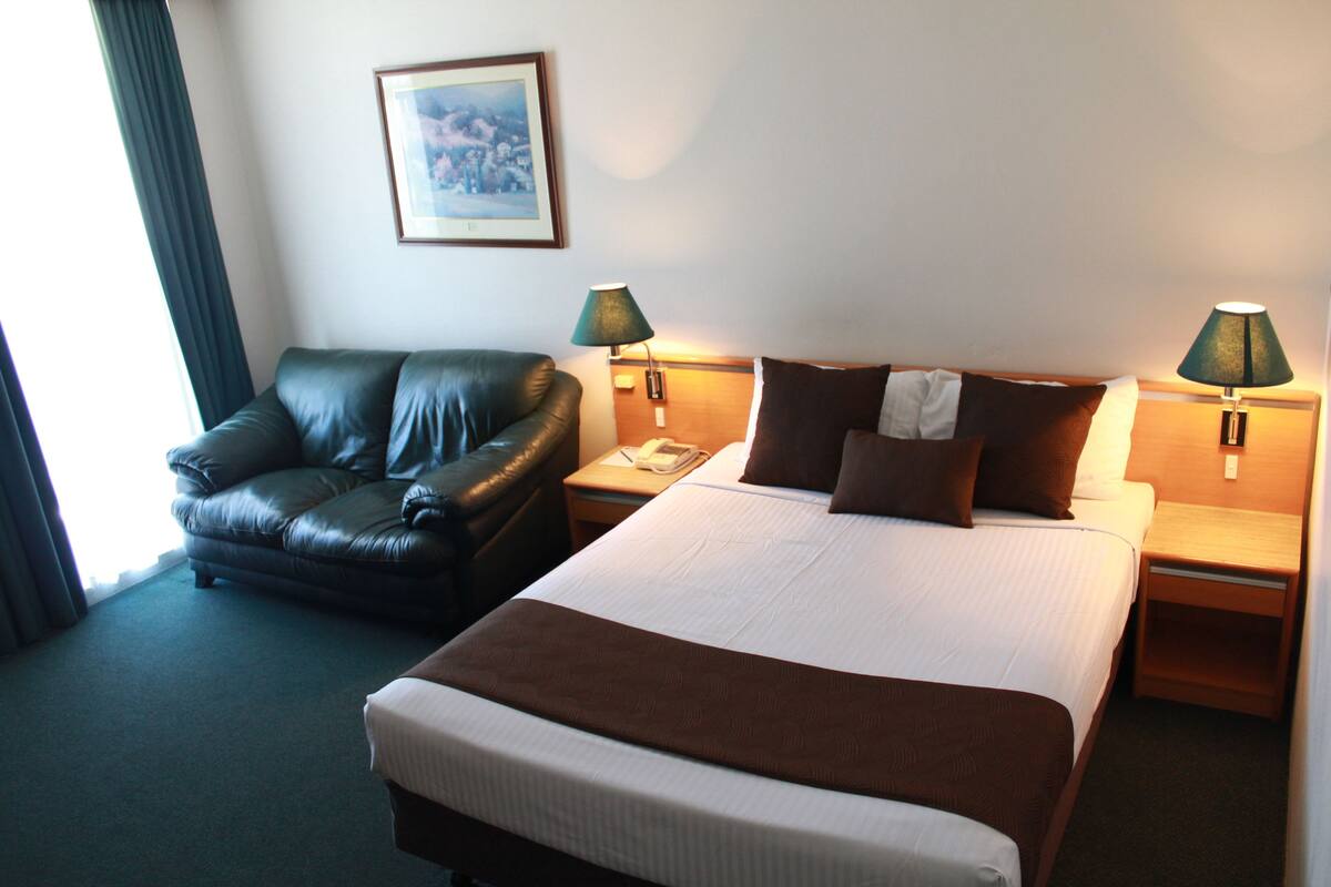 A hotel room with a bed, a leather armchair, two bedside lamps, a framed picture on the wall, and a window with curtains.