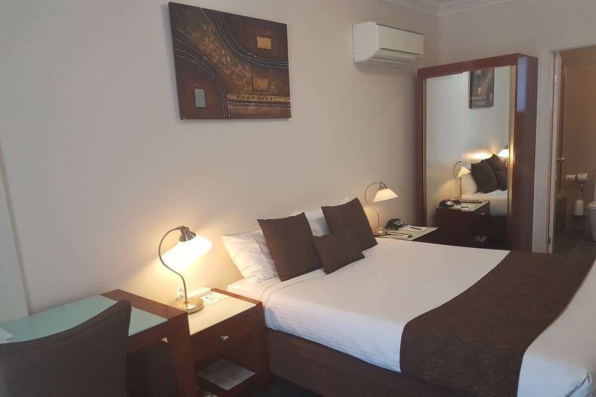 A hotel room with a large bed, two bedside tables with lamps, a wall-mounted air conditioner, and a painting above the bed.