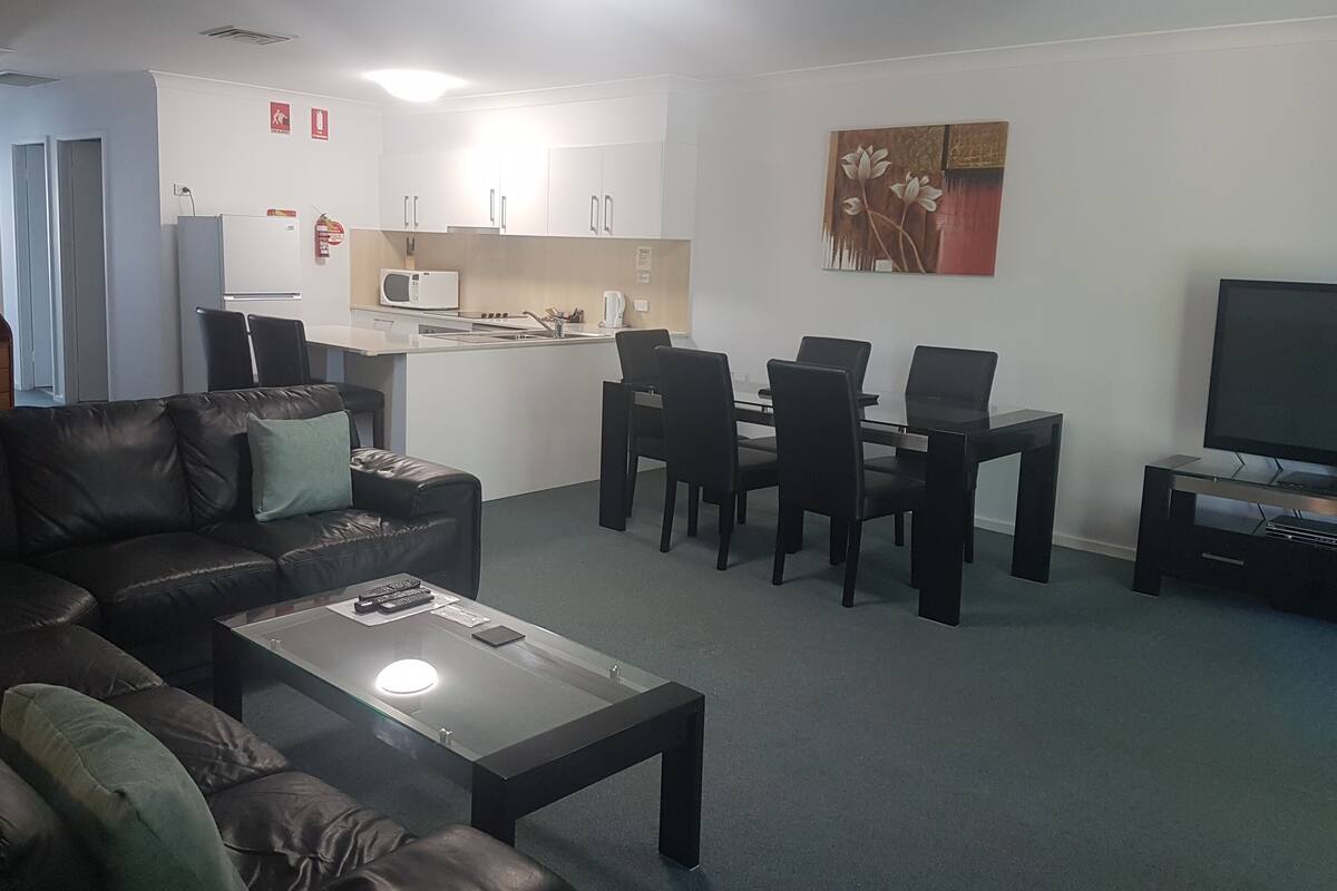 A living room with a sofa, coffee table, TV, and a dining area with chairs and a table. There is a kitchen area with cabinets and a microwave.
