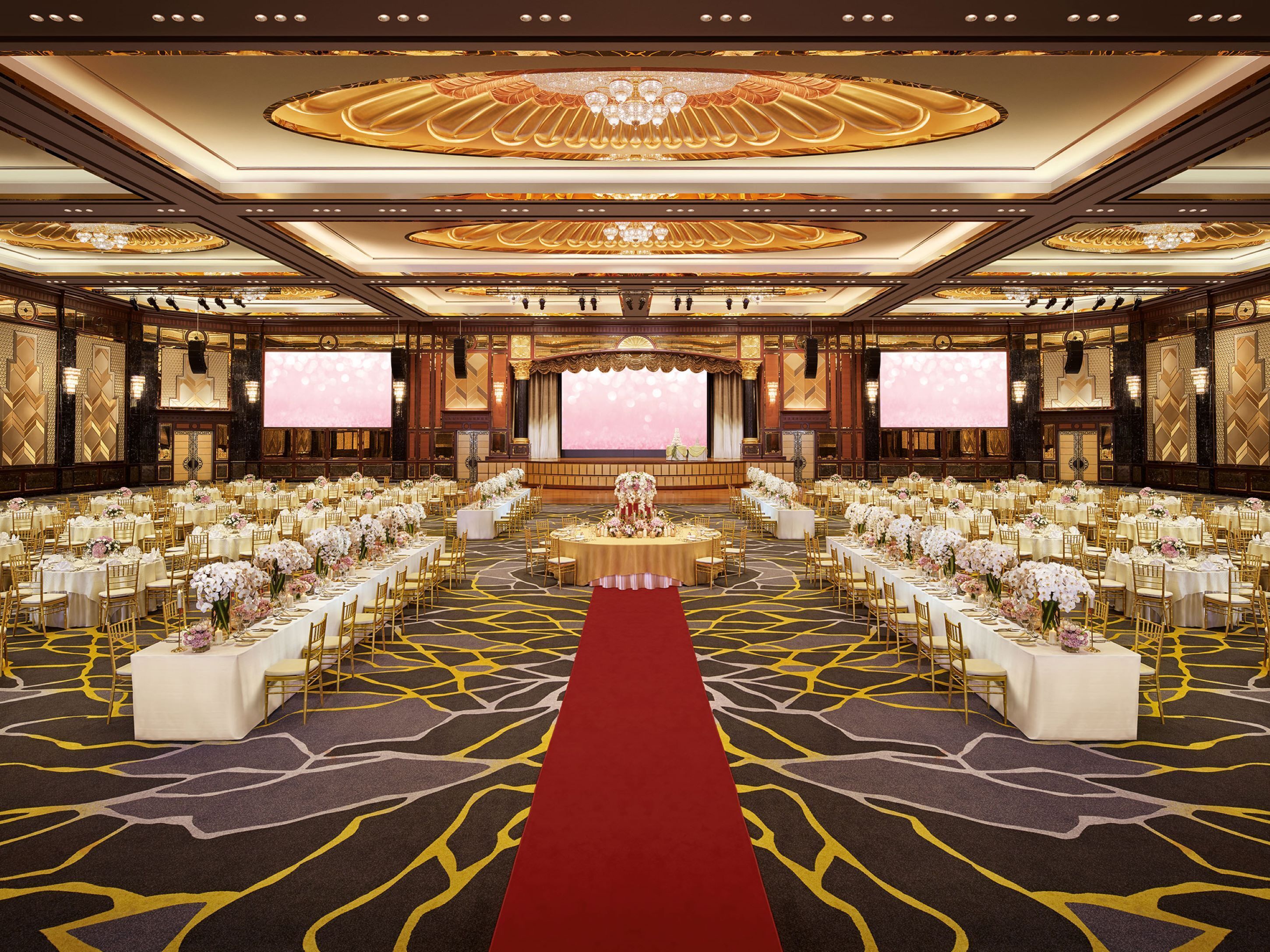 A grand ballroom with a red carpet, multiple round tables, and a stage with a large screen.