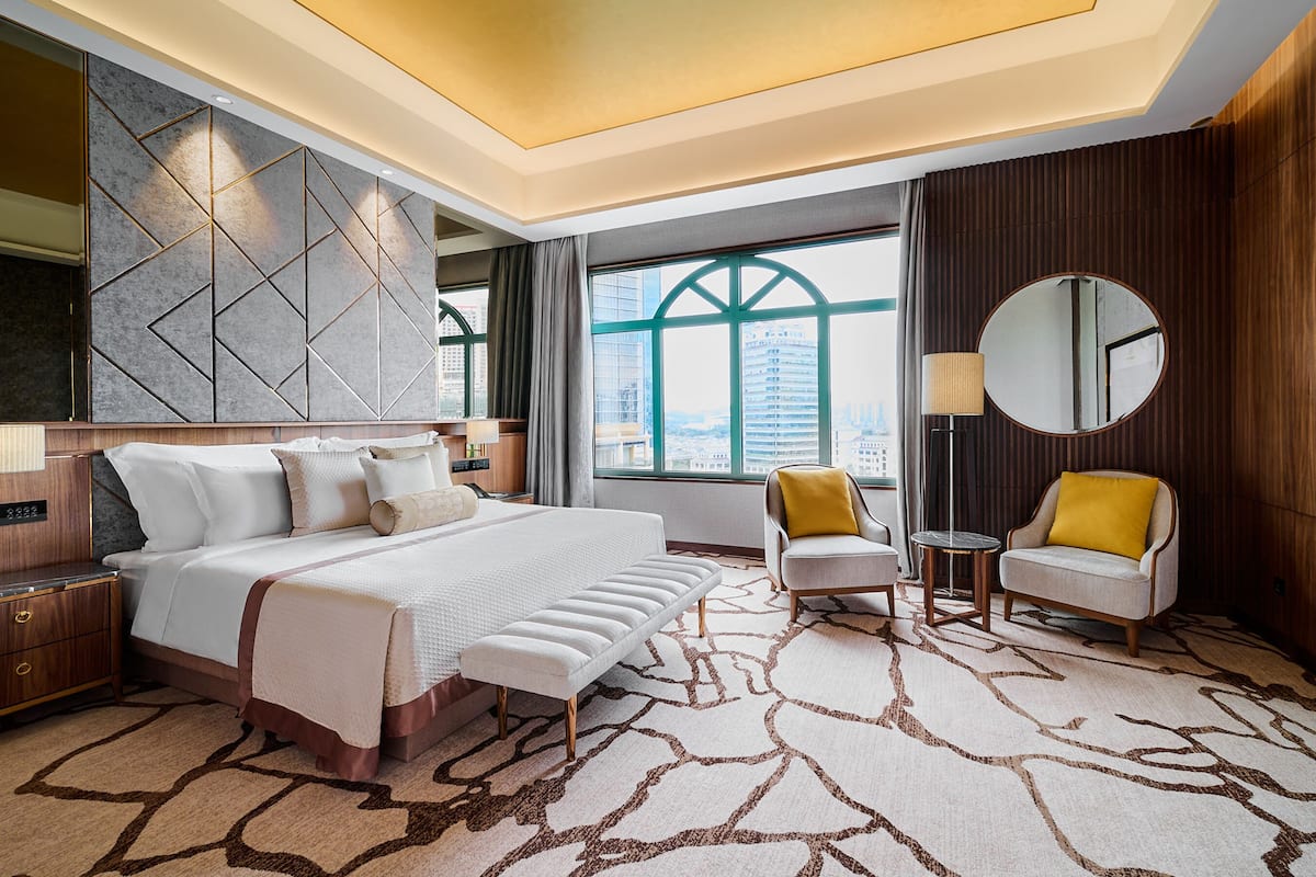 A modern hotel room with a large bed, a seating area with two chairs, a round mirror, and a view of the city through large windows.
