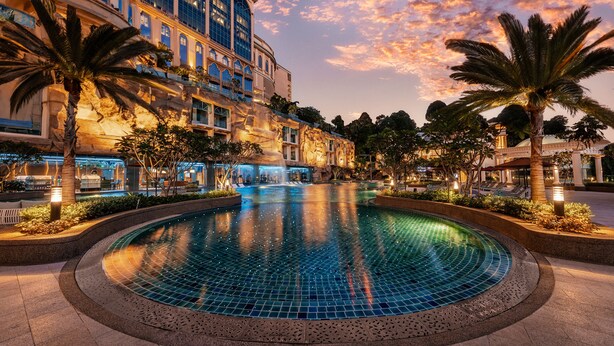 Un hotel con una amplia piscina exterior rodeada de palmeras y un cielo al atardecer.
