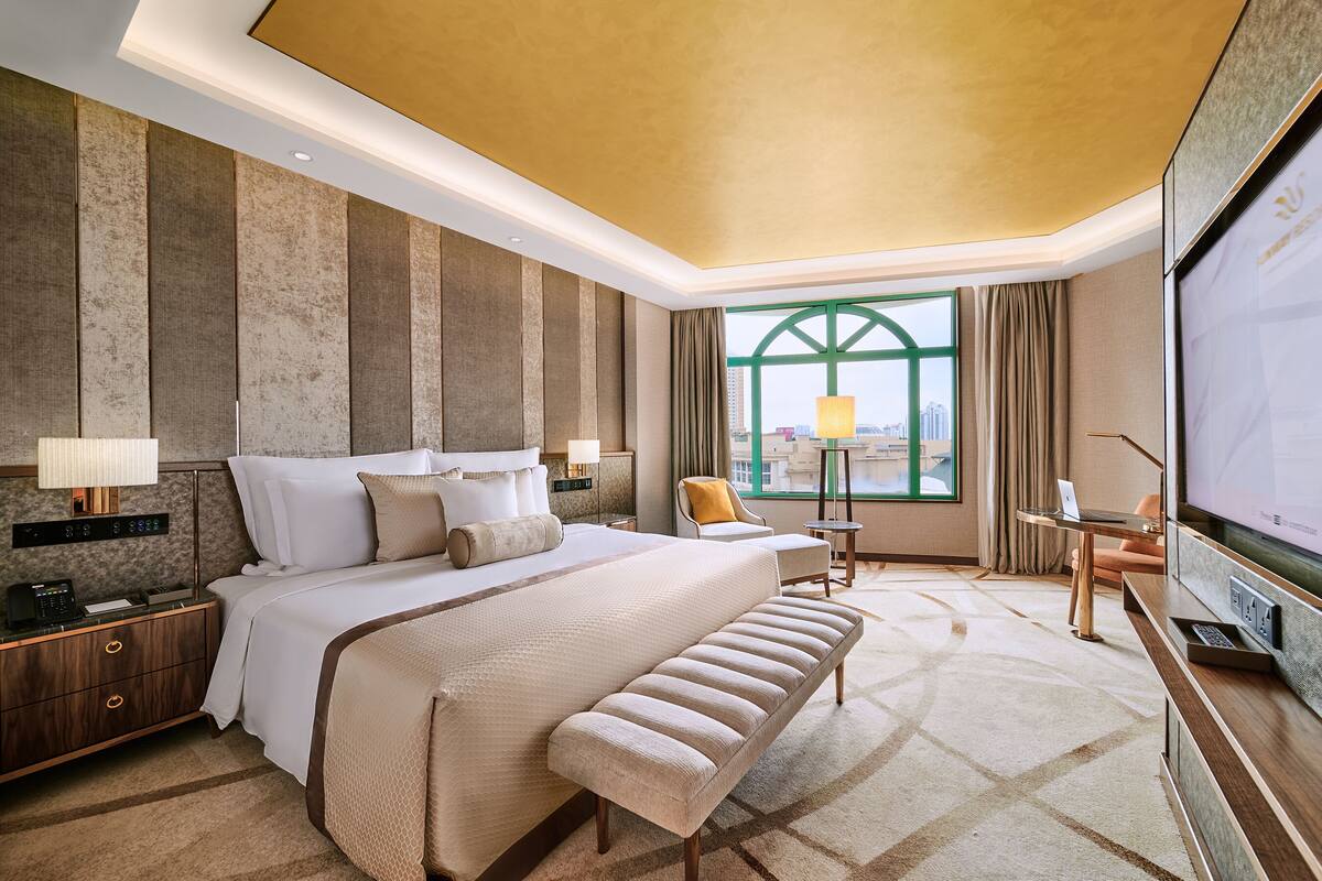 A hotel room with a large bed, a desk with a laptop, a TV, and a view of the city through a large window.