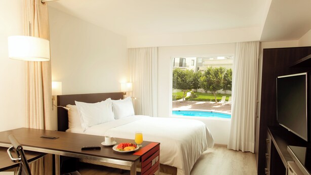 A hotel room with a bed, a desk with a chair, a television, and a window with a view of a swimming pool and greenery. - Fiesta Inn Express Monclova (Monclova)