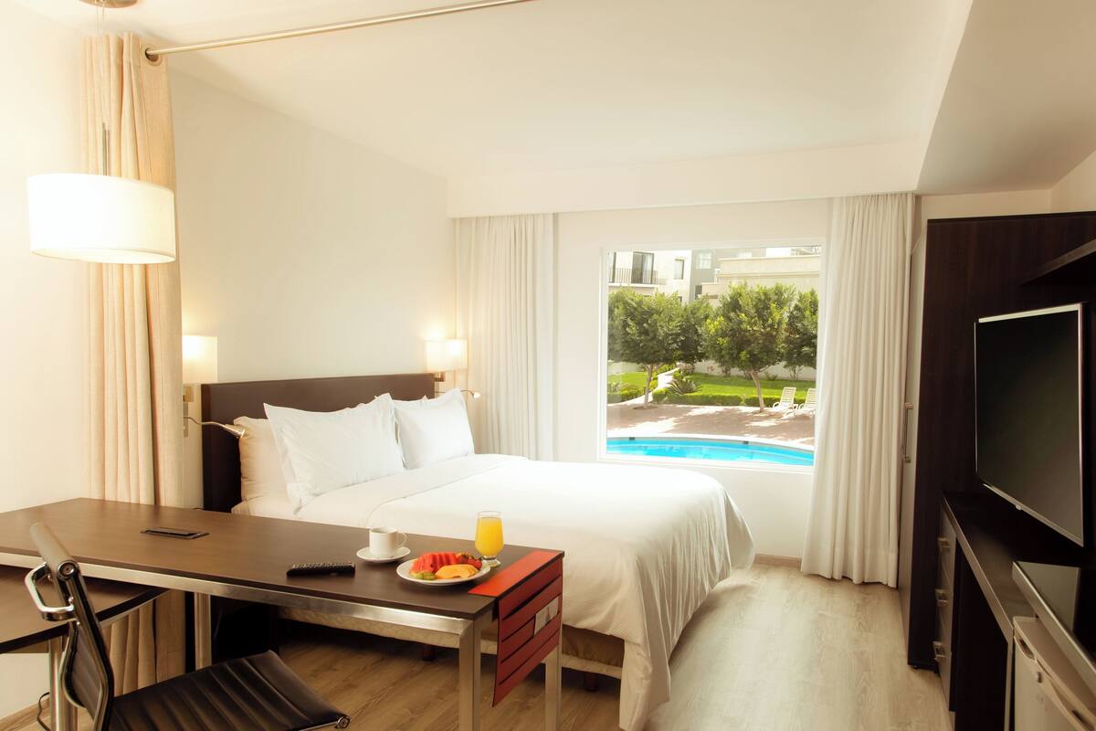 Une chambre d’hôtel comprenant un lit, un bureau avec une chaise, une télévision et une fenêtre donnant sur une piscine et un espace verdoyant.