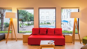 A red sofa with a magazine on a small table, flanked by two floor lamps and two windows with views of a pool and buildings. - Fiesta Inn Express Monclova (Monclova)