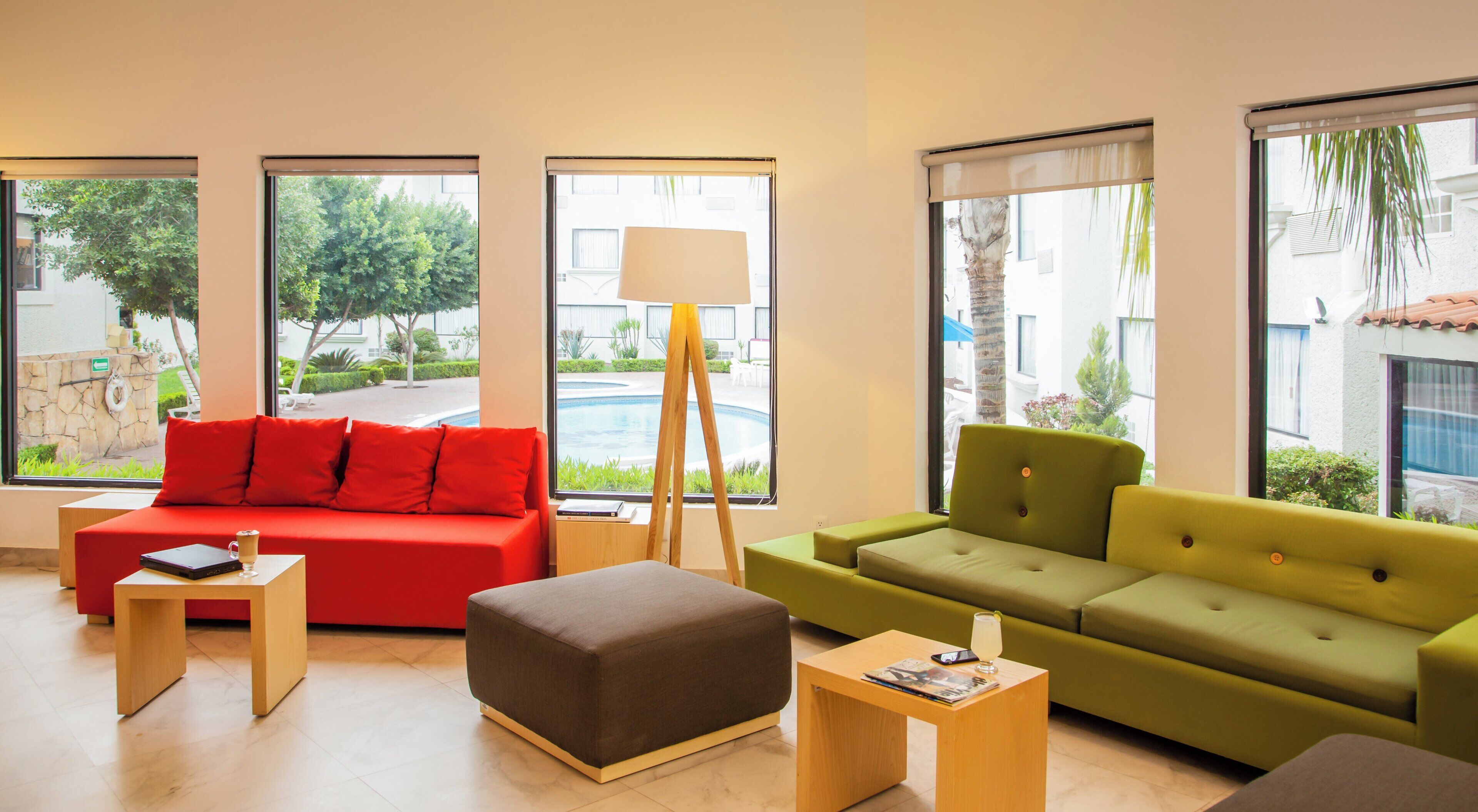 A modern living room with a red sofa, a green sofa, a brown ottoman, and a floor lamp. Large windows provide views of a pool and greenery.