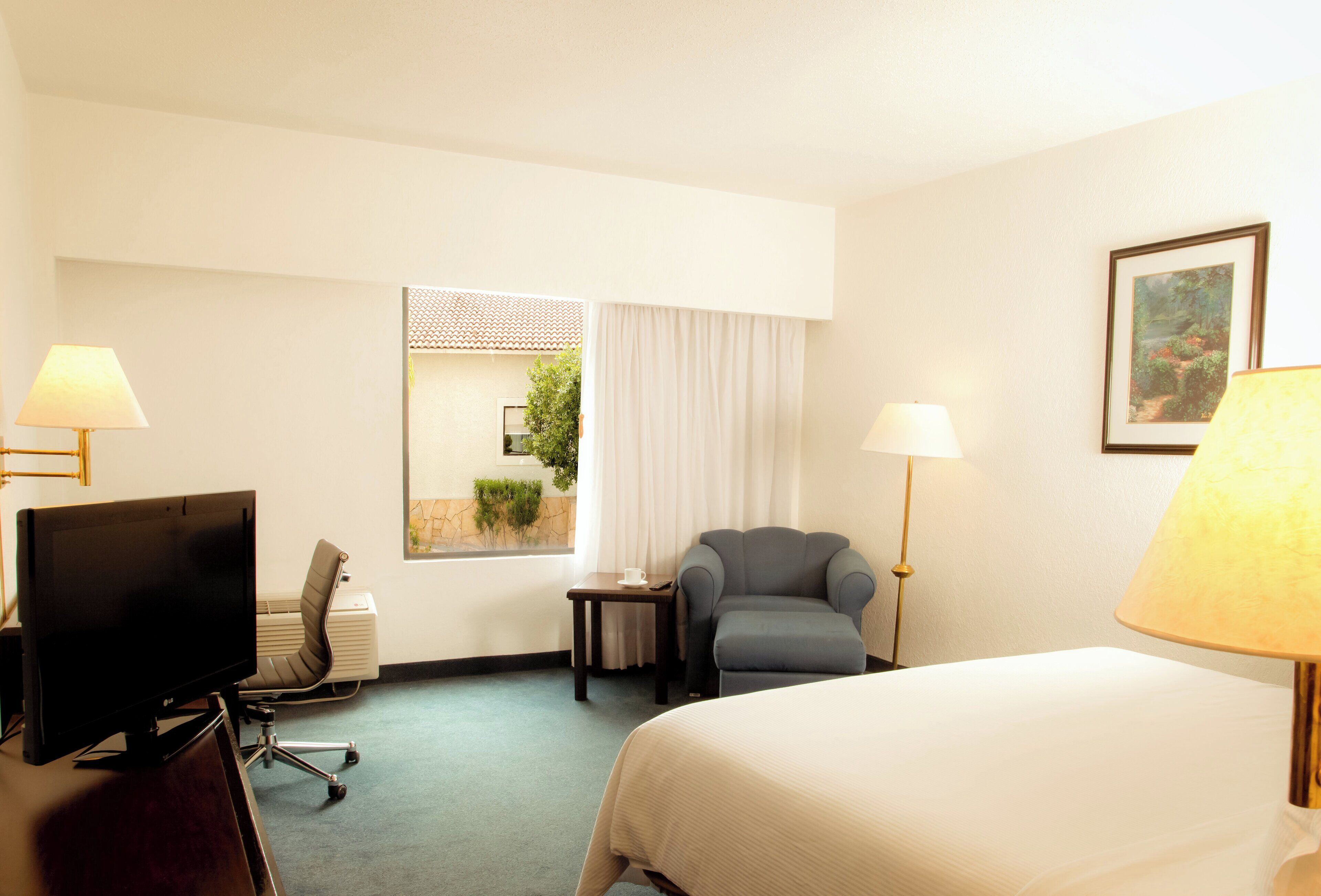 A hotel room with a bed, a desk with a computer, a chair, a lamp, and a window with a view of greenery.