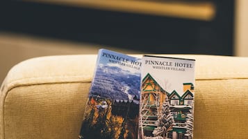 Two boxes of Pinnacle Hotel Whistler Village chocolate on a beige couch with a blurred fireplace in the background.