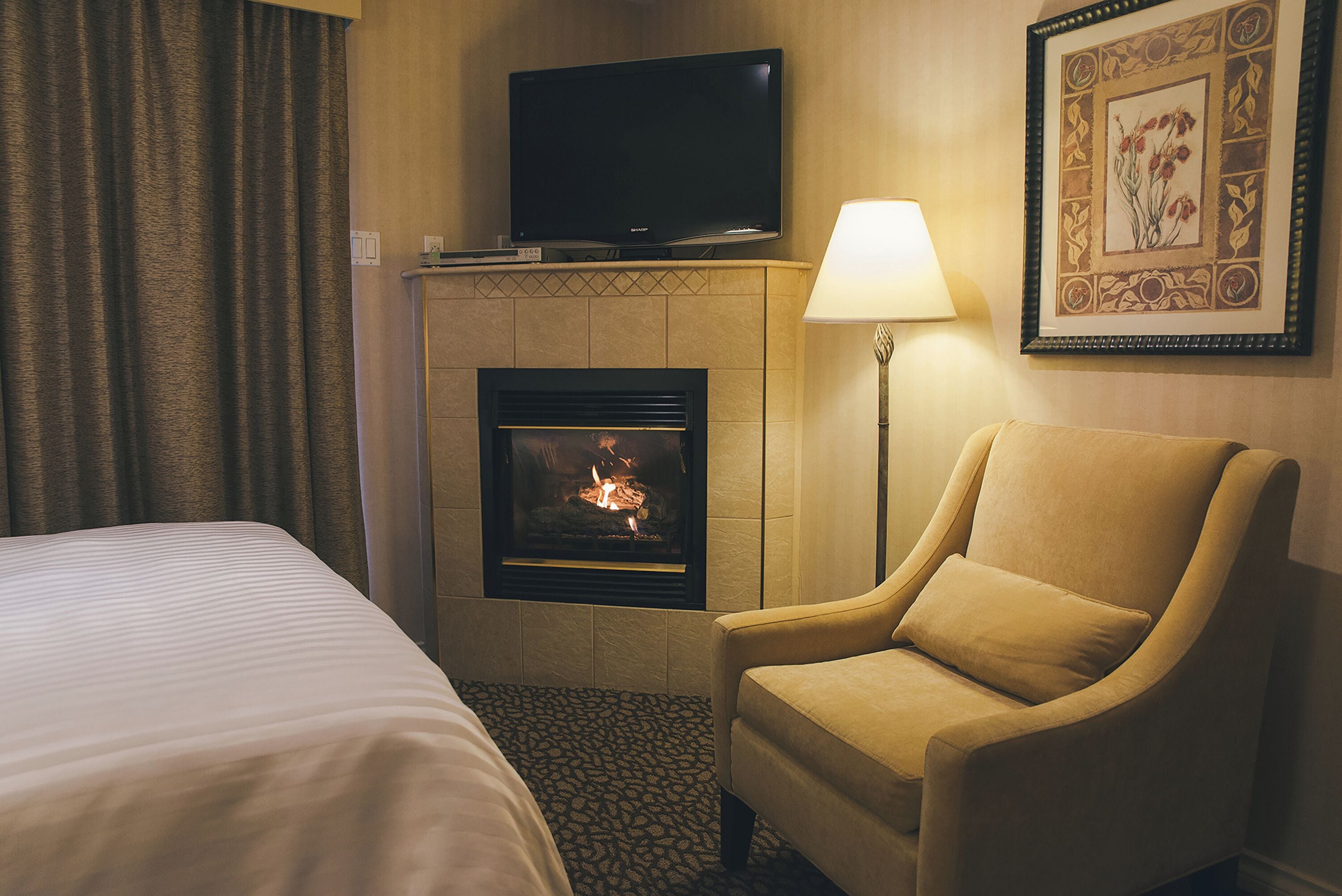 A hotel room with a bed, a chair, a fireplace, a TV, and a lamp.