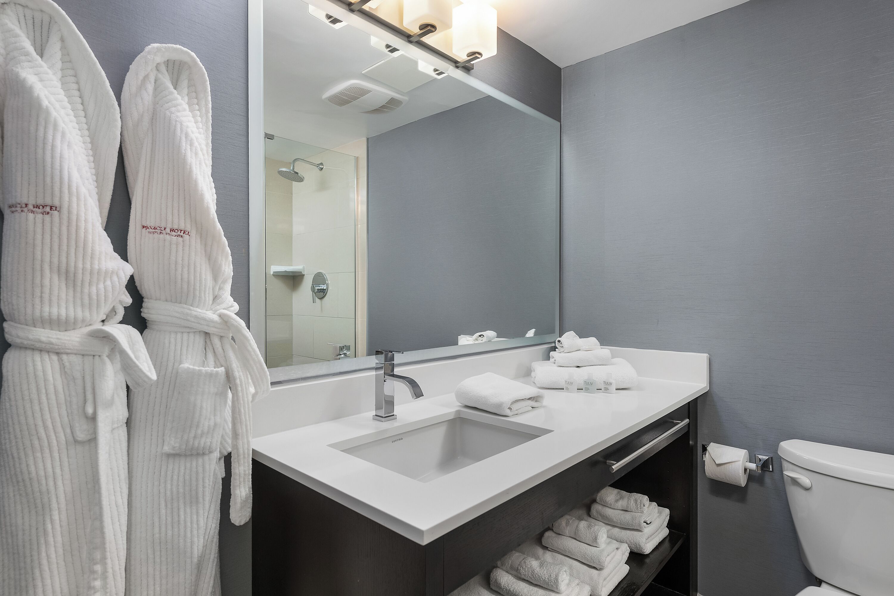 Un baño moderno con un espejo grande, un lavamanos y un inodoro. Hay batas blancas colgadas en la pared y un estante con toallas dobladas.