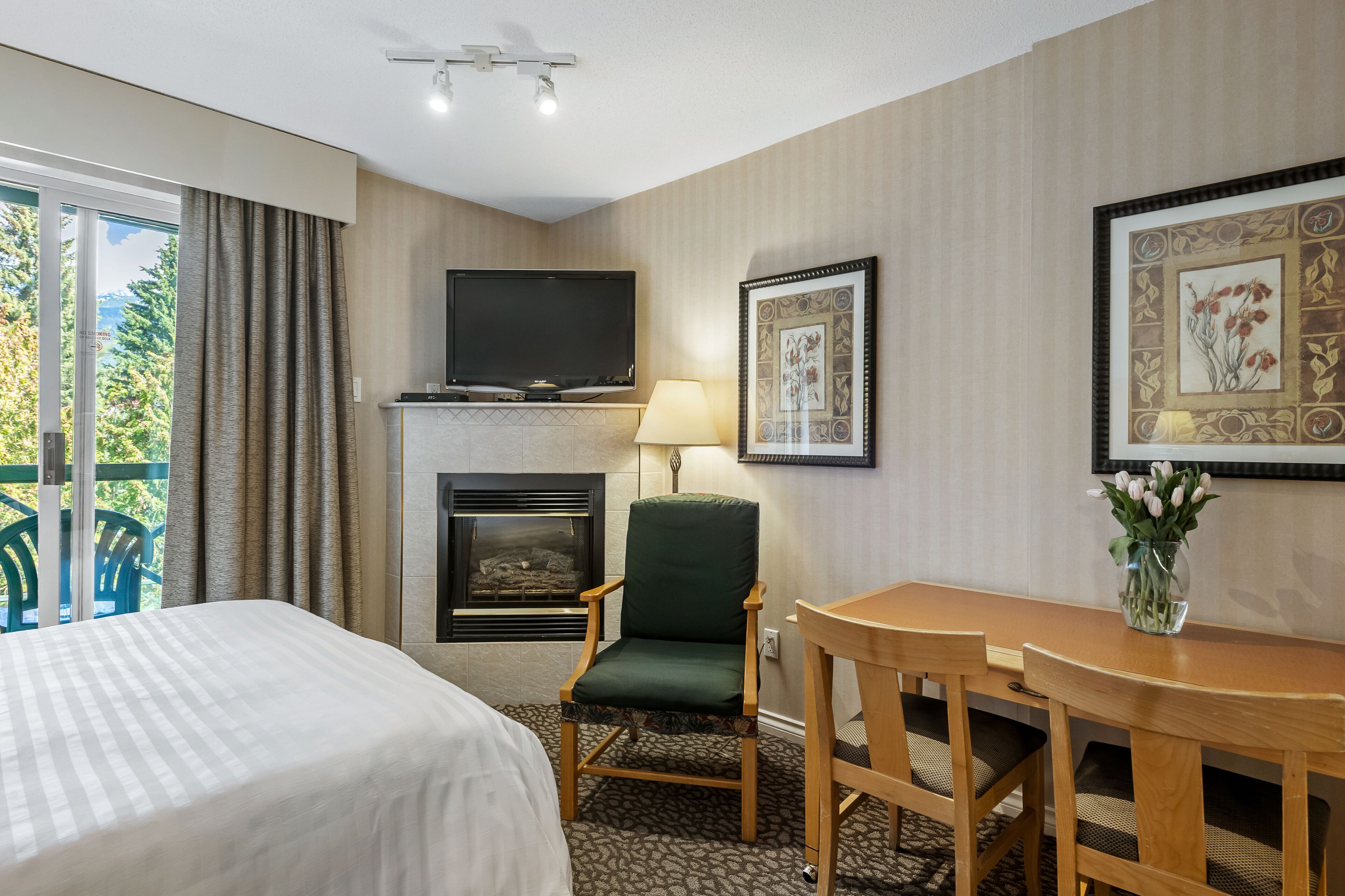 A hotel room with a bed, a chair, a table, a fireplace, a TV, and a window with curtains.