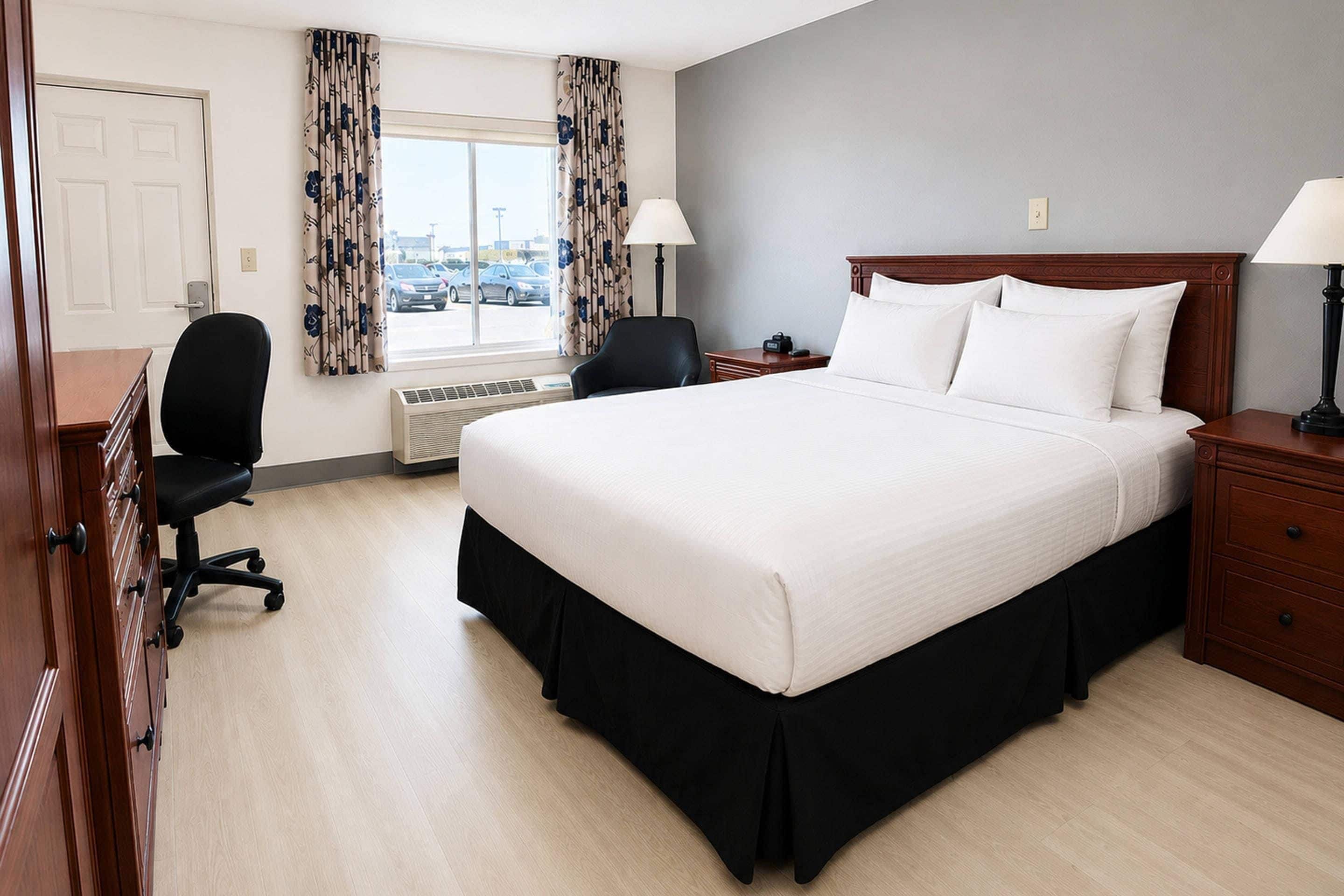 A hotel room with a large bed, a desk with a chair, a window with curtains, and a nightstand with a lamp.