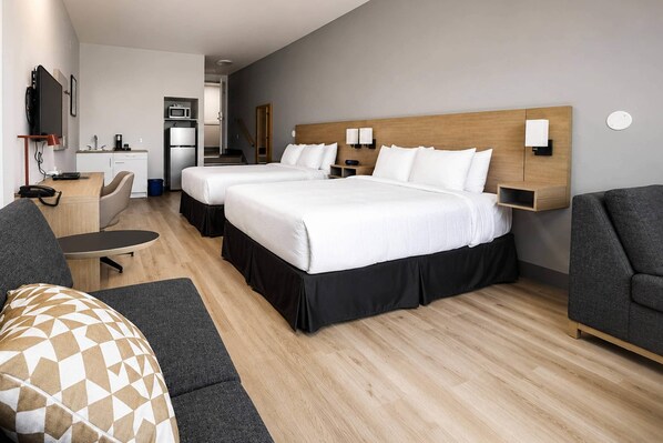 A hotel room with two beds, a desk, a chair, a sofa, a TV, and a kitchenette.