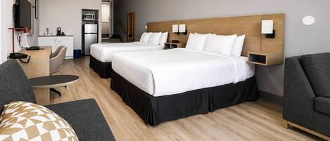 A hotel room with two beds, a desk, a chair, a sofa, a TV, and a kitchenette.