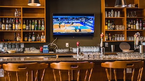 Zona de bar con una televisión mostrando un partido de baloncesto, estanterías con diferentes botellas, vasos y una barra con taburetes.