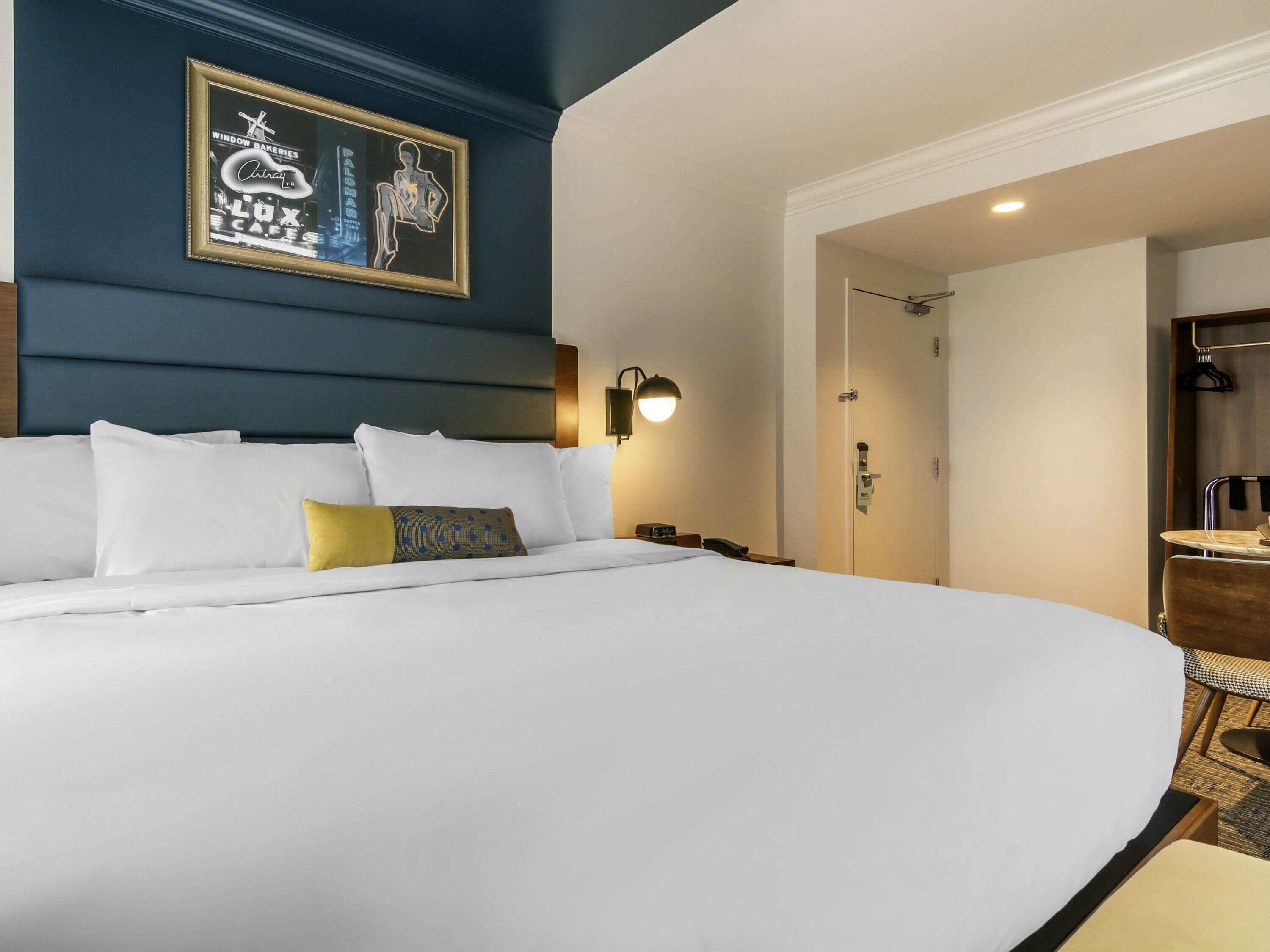 Une chambre d’hôtel avec un grand lit, une œuvre d’art encadrée au mur, une lampe de chevet et une porte donnant sur une salle de bain.
